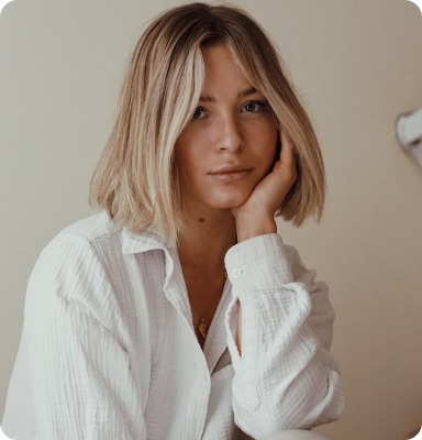 A portrait of a woman with shoulder-length blonde hair, resting her cheek on her hand, wearing a white button-up shirt. She has a pensive expression and is looking slightly away from the camera.